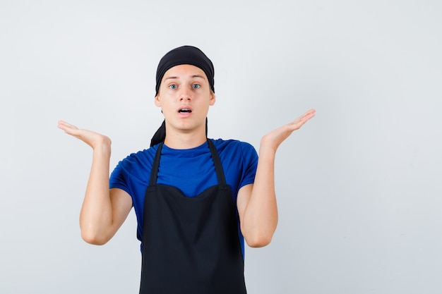 Hombre joven cocinero fingiendo sostener algo en camiseta, delantal y mirando perplejo, vista frontal.