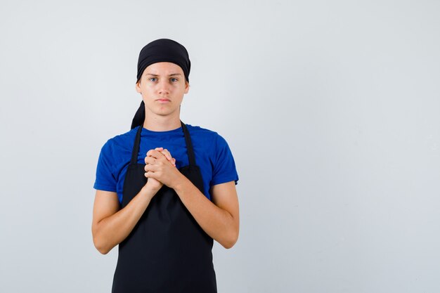 Hombre joven cocinero mostrando las manos juntas en gesto de súplica en camiseta, delantal y mirando esperanzado, vista frontal.