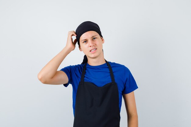 Hombre joven cocinero rascándose la cabeza en camiseta, delantal y mirando pensativo. vista frontal.