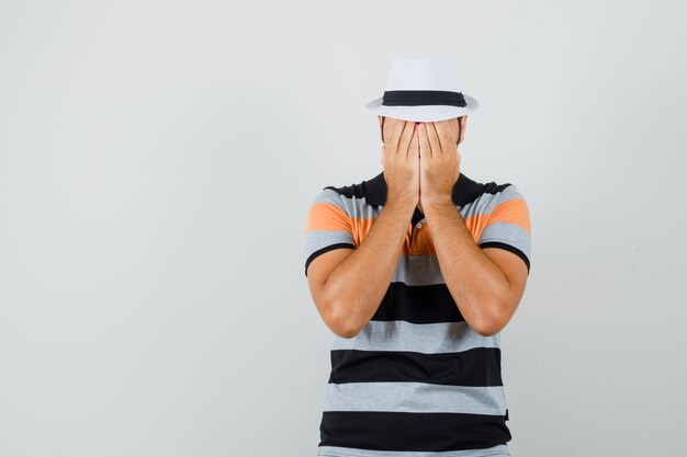 Hombre joven cogidos de la mano en la cara con camiseta a rayas, sombrero y mirando triste. espacio para texto