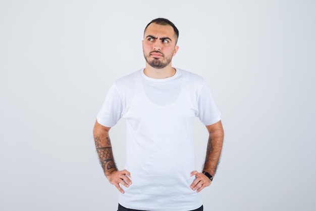 Hombre joven cogidos de la mano en la cintura con camiseta blanca y pantalón negro y mirando pensativo