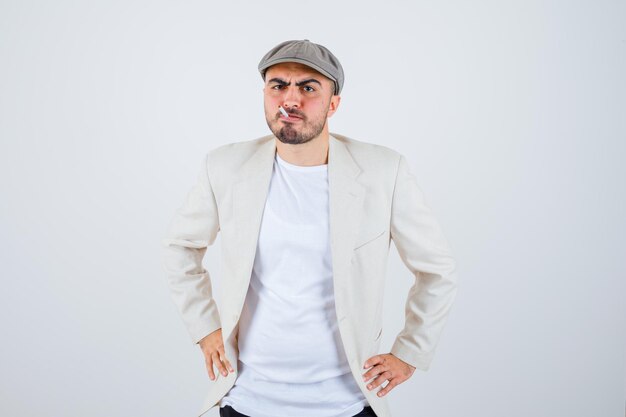 Hombre joven cogidos de la mano en la cintura mientras fuma en camiseta blanca, chaqueta y gorra gris y mirando enojado