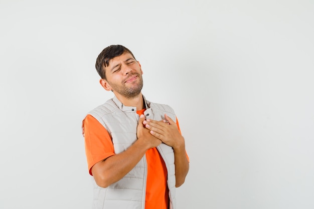 Hombre joven cogidos de la mano en el corazón en camiseta, chaqueta y mirando incómodo, vista frontal.