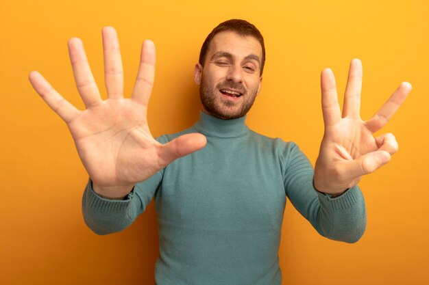 Hombre joven complacido mirando al frente mostrando ocho con las manos guiñando un ojo aislado en la pared naranja