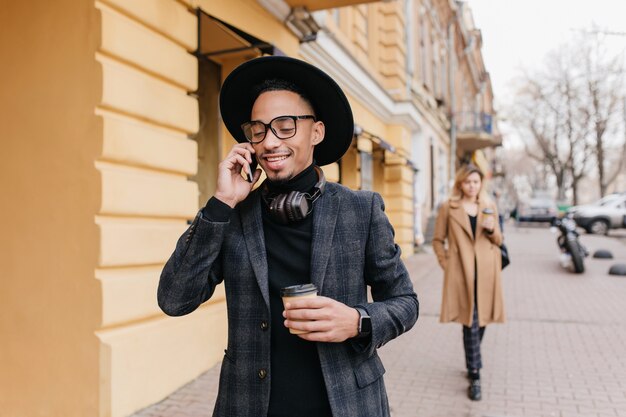Hombre joven complacido con piel morena tomando café en la calle. Chico africano feliz con taza de café con leche llamando a alguien.