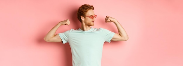 Foto gratuita hombre joven confiado con cabello rojo con gafas de sol de verano y camiseta que muestra un cuerpo fuerte y en forma m