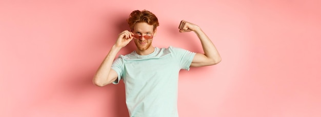 Hombre joven confiado con cabello rojo con gafas de sol de verano y camiseta que muestra un cuerpo fuerte y en forma m