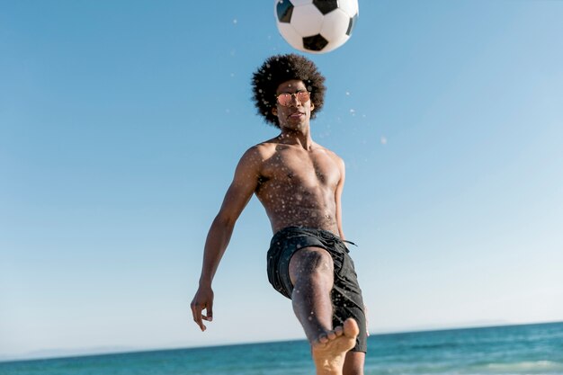 Hombre joven confiado pateando la pelota en la orilla del mar