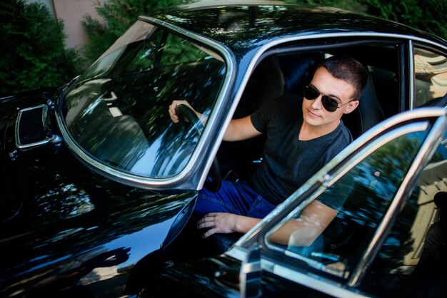 El hombre joven confidente en gafas de sol se sienta dentro de un coche retro del deporte negro