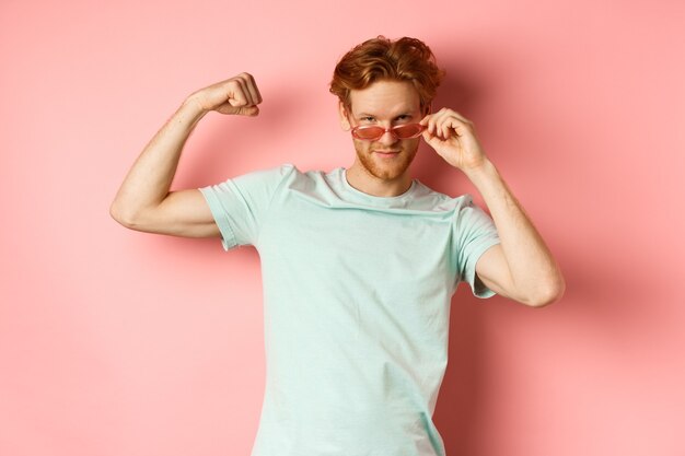 Hombre joven confidente con el pelo rojo con gafas de sol de verano y camiseta mostrando cuerpo fuerte y en forma ...