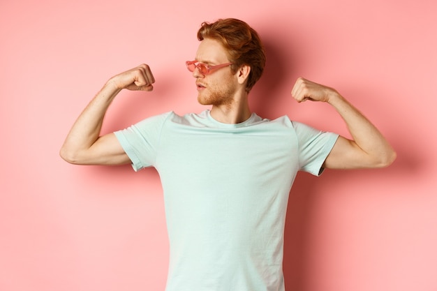 Hombre joven confidente con el pelo rojo con gafas de sol de verano y camiseta mostrando cuerpo fuerte y en forma ...