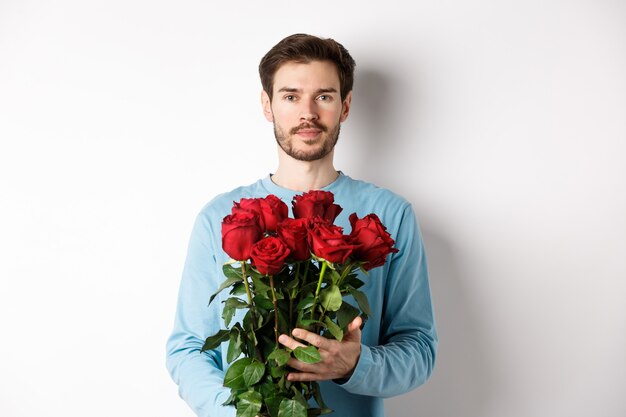 Hombre joven confidente traer flores en la fecha del día de San Valentín, sosteniendo un ramo romántico, de pie sobre fondo blanco