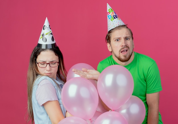 Hombre joven confundido con sombrero de fiesta apunta a la niña ofendida de pie con globos de helio aislados en la pared rosa