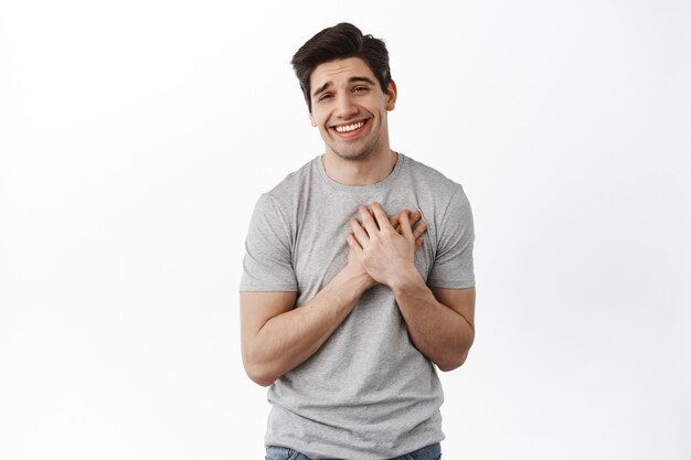 Hombre joven conmovido y halagado cogidos de la mano en el corazón y sonriendo, sentirse agradecido, agradeciendo y luciendo sincero, de pie en camiseta contra la pared blanca