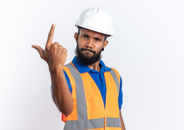 Hombre joven constructor afroamericano molesto en uniforme con casco de seguridad apuntando hacia arriba aislado en la pared blanca con espacio de copia