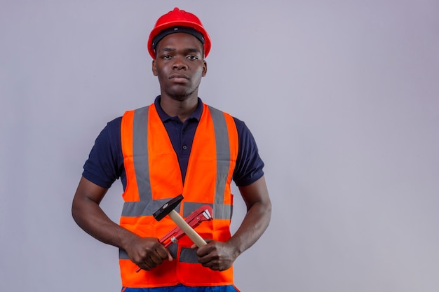 Hombre joven constructor afroamericano vestido con chaleco de construcción y casco de seguridad con llave ajustable y martillo en forma de cruz con cara seria de pie