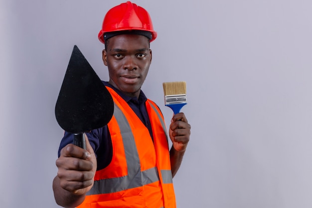 Hombre joven constructor afroamericano vestido con chaleco de construcción y casco de seguridad mostrando espátula y sosteniendo pincel mirando con sonrisa confiada en blanco aislado
