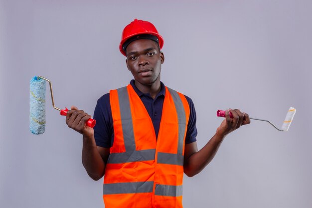 Hombre joven constructor afroamericano vestido con chaleco de construcción y casco de seguridad con rodillo de pintura y pincel de pie con expresión confusa con los brazos y las manos levantadas