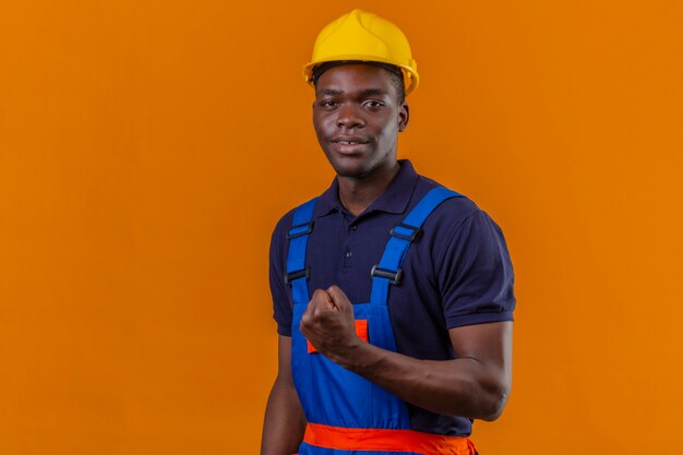 Hombre joven constructor afroamericano vestido con uniforme de construcción y casco de seguridad mostrando puño sonriente de pie con cara feliz celebrando el concepto ganador de la victoria en naranja aislado