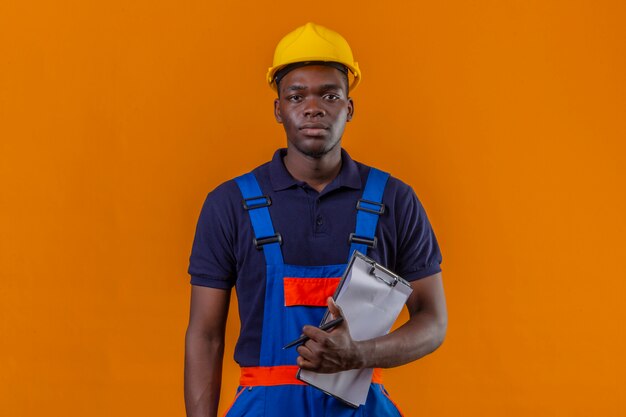 Hombre joven constructor afroamericano vestido con uniforme de construcción y casco de seguridad de pie con portapapeles y bolígrafo en mano con expresión seria y segura en naranja aislado