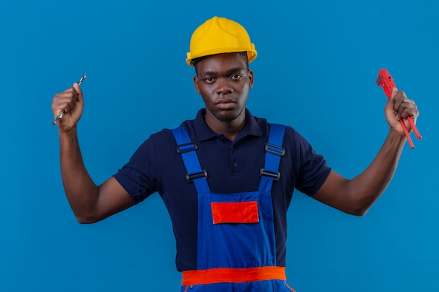 Hombre joven constructor afroamericano vestido con uniforme de construcción y casco de seguridad sosteniendo llaves ajustables en la mano levantada con expresión enojada agresiva de pie