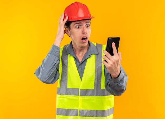 Hombre joven constructor asustado en uniforme sosteniendo y mirando el teléfono poniendo la mano en la cabeza aislada en la pared amarilla