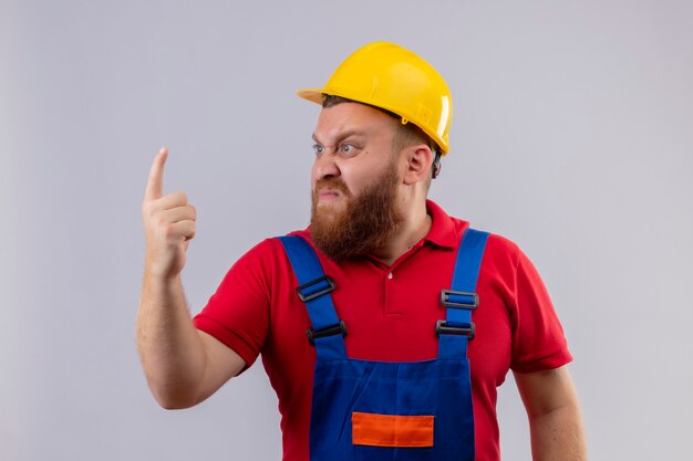 Hombre joven constructor barbudo molesto en uniforme de construcción y casco de seguridad mirando a un lado con cara enojada apuntando con el dedo hacia arriba