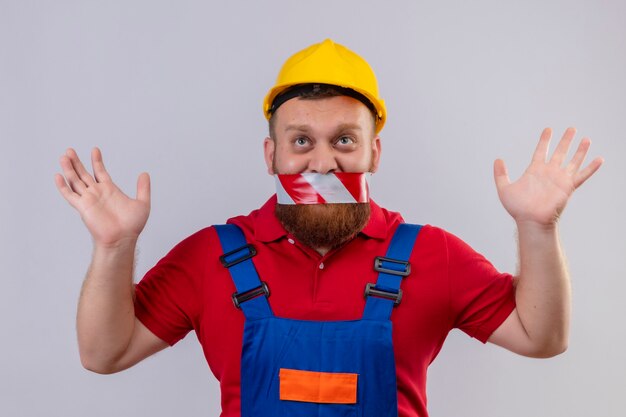 Hombre joven constructor barbudo en uniforme de construcción y casco de seguridad con cinta adhesiva sobre la boca mirando hacia arriba levantando las manos en sarrender