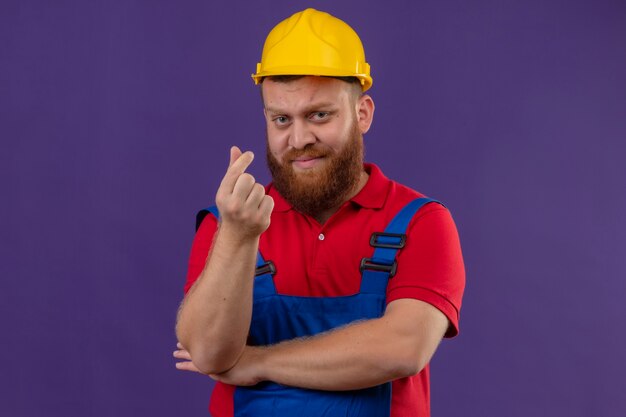 Hombre joven constructor barbudo en uniforme de construcción y casco de seguridad disgustado haciendo gesto de dinero con la mano frotando los dedos pidiendo dinero sobre fondo púrpura