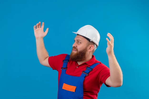 Hombre joven constructor barbudo en uniforme de construcción y casco de seguridad encogiéndose de hombros, luciendo confuso e incierto, sin respuesta, extendiendo las palmas