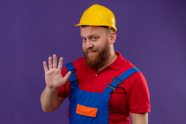 Hombre joven constructor barbudo en uniforme de construcción y casco de seguridad con expresión escéptica mostrando y apuntando hacia arriba con los dedos número cinco sobre fondo púrpura