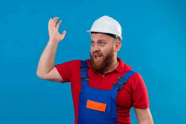 Hombre joven constructor barbudo en uniforme de construcción y casco de seguridad gritando mirando confundido levantando la mano