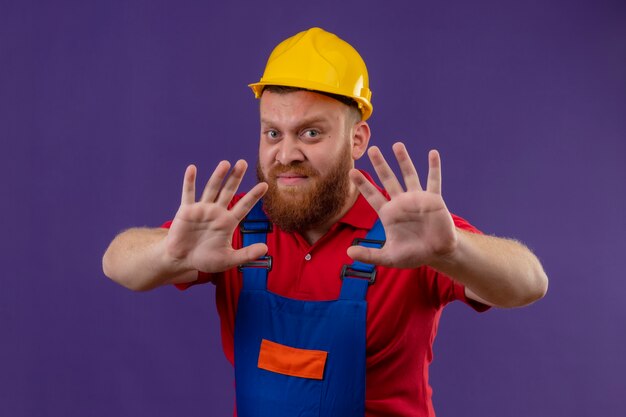 Hombre joven constructor barbudo en uniforme de construcción y casco de seguridad haciendo gesto de defensa de rechazo con las manos abiertas, con expresión de disgusto sobre fondo púrpura