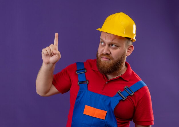 Hombre joven constructor barbudo en uniforme de construcción y casco de seguridad mirando hacia arriba con el ceño fruncido apuntando el dedo hacia arriba sobre fondo púrpura