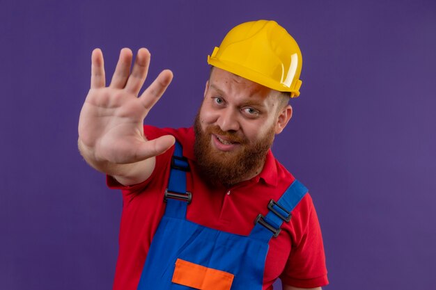 Hombre joven constructor barbudo en uniforme de construcción y casco de seguridad mirando asustado con la mano abierta haciendo señal de stop sobre fondo púrpura