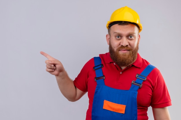 Hombre joven constructor barbudo en uniforme de construcción y casco de seguridad mirando a la cámara apuntando con el dedo hacia el lado con cara seria