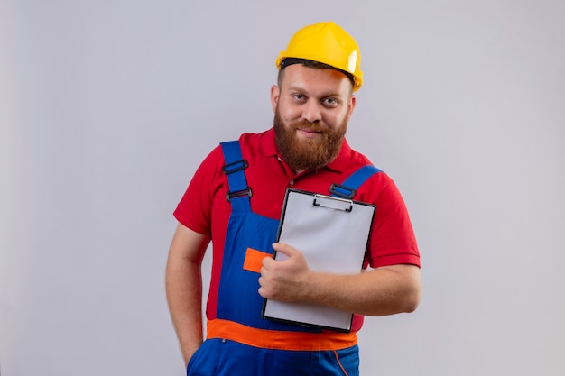 Foto gratuita hombre joven constructor barbudo en uniforme de construcción y casco de seguridad mirando a cámara sosteniendo portapapeles sonriendo confiado