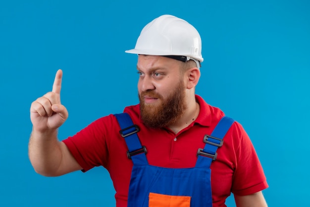 Hombre joven constructor barbudo en uniforme de construcción y casco de seguridad mirando a un lado confundido dedo apuntando hacia arriba