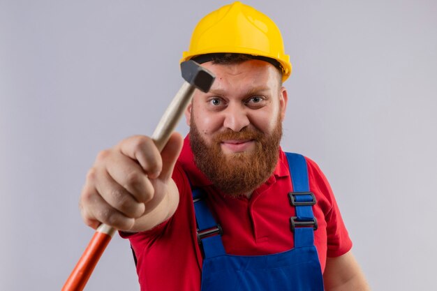 Hombre joven constructor barbudo en uniforme de construcción y casco de seguridad mostrando el martillo a la cámara mirando a la cámara positiva y feliz