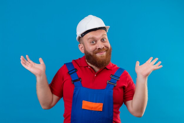 Hombre joven constructor barbudo en uniforme de construcción y casco de seguridad con las palmas levantadas, encogiéndose de hombros sonriendo, sin tener respuesta