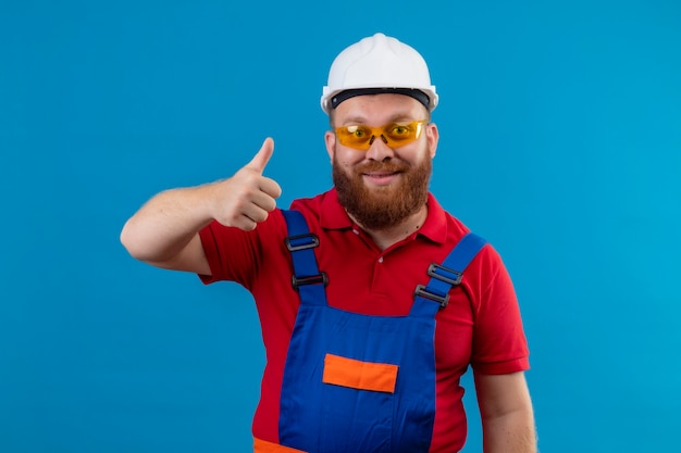 Hombre joven constructor barbudo en uniforme de construcción y casco de seguridad positivo y feliz mostrando los pulgares um sonriendo amable