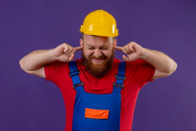 Hombre joven constructor barbudo en uniforme de construcción y casco de seguridad que cubre las orejas con los dedos con expresión molesta por el ruido del sonido fuerte sobre fondo púrpura