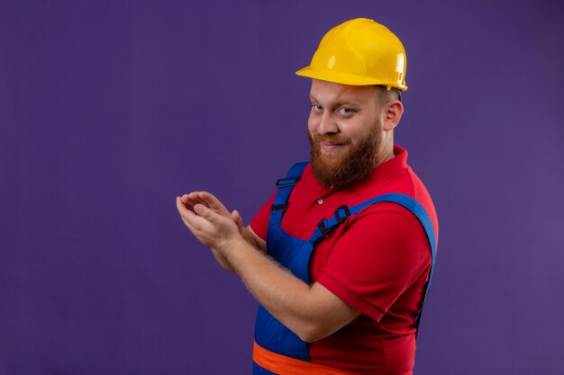 Hombre joven constructor barbudo en uniforme de construcción y casco de seguridad que parece seguro con una sonrisa en la cara aplaudiendo sobre fondo púrpura