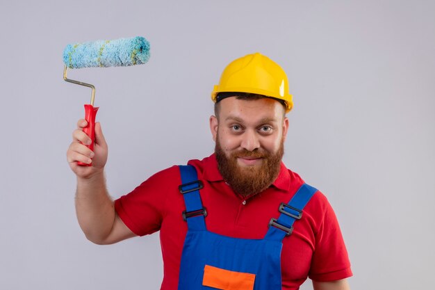 Hombre joven constructor barbudo en uniforme de construcción y casco de seguridad con rodillo de pintura mirando a cámara sonriendo feliz y positivo