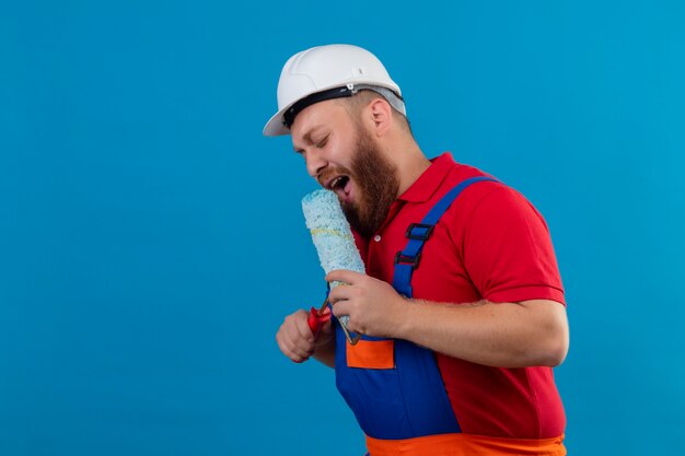 Hombre joven constructor barbudo en uniforme de construcción y casco de seguridad con rodillo de pintura usando como micrófono cantando, divirtiéndose en el trabajo