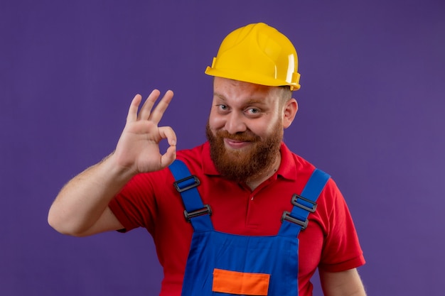 Hombre joven constructor barbudo en uniforme de construcción y casco de seguridad sonriendo alegremente haciendo bien firmar sobre fondo púrpura