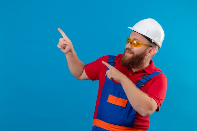 Hombre joven constructor barbudo en uniforme de construcción y casco de seguridad sonriendo feliz y positivo apuntando con los dedos hacia el lado