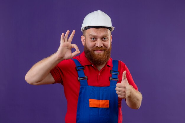 Hombre joven constructor barbudo en uniforme de construcción y casco de seguridad sonriendo feliz y positivo haciendo bien cantar mostrando los pulgares hacia arriba sobre fondo púrpura