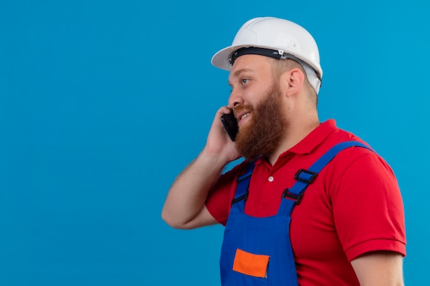 Foto gratuita hombre joven constructor barbudo en uniforme de construcción y casco de seguridad sonriendo hablando por teléfono móvil