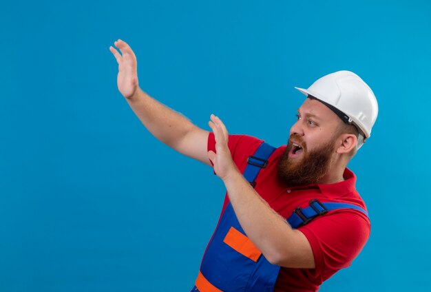 Hombre joven constructor barbudo en uniforme de construcción y casco de seguridad tomados de la mano haciendo gesto de defensa mirando asustado gritando